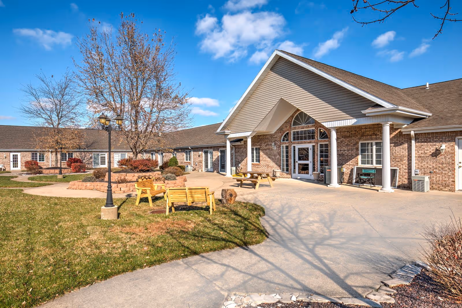 Exterior view of a senior living facility with a brick facade and a peaked roof entrance. There is a paved walkway leading to the entrance, surrounded by green grass, leafless trees, a lamppost, wooden benches, and a picnic table under a clear blue sky.