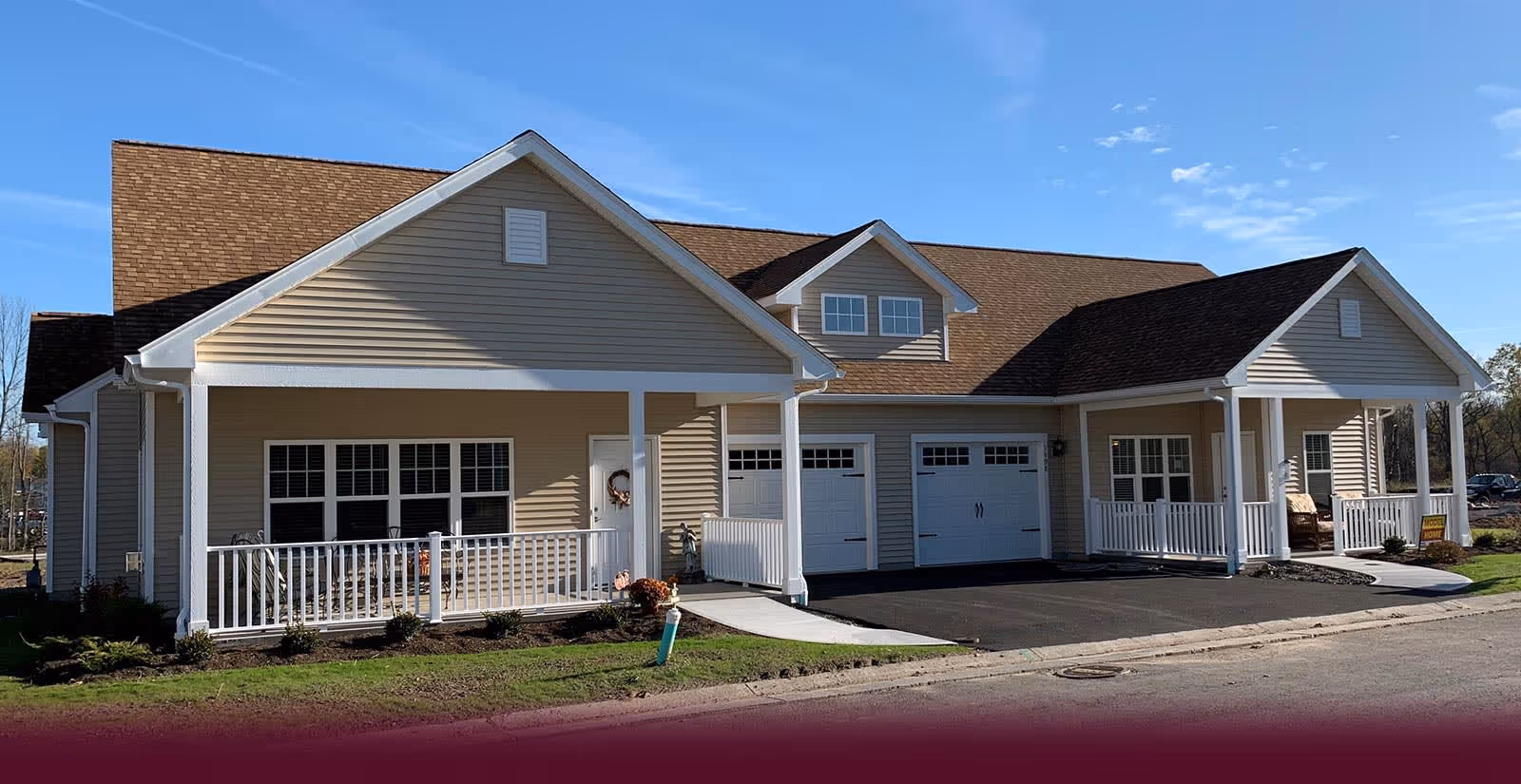 Exterior view of a single-story senior living community building with beige siding, white trim, two garage doors, a covered porch with white railings, and a paved driveway under a clear blue sky.