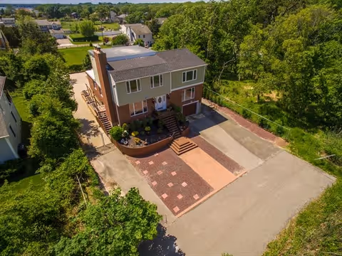 Aerial view of a two-story residential building with a brick and beige exterior, surrounded by greenery and trees. The building has a driveway and a paved area in front, with steps leading up to the main entrance. Other houses and a road are visible in the background.