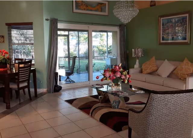 A cozy living room with a beige sofa adorned with yellow and white pillows, a glass coffee table with a flower arrangement, and a patterned armchair. The room has green walls, a chandelier, framed artwork, and large sliding glass doors leading to an outdoor patio with a chair and a fenced yard. To the left, there is a dining area with a wooden table and chairs near a window with blinds.
