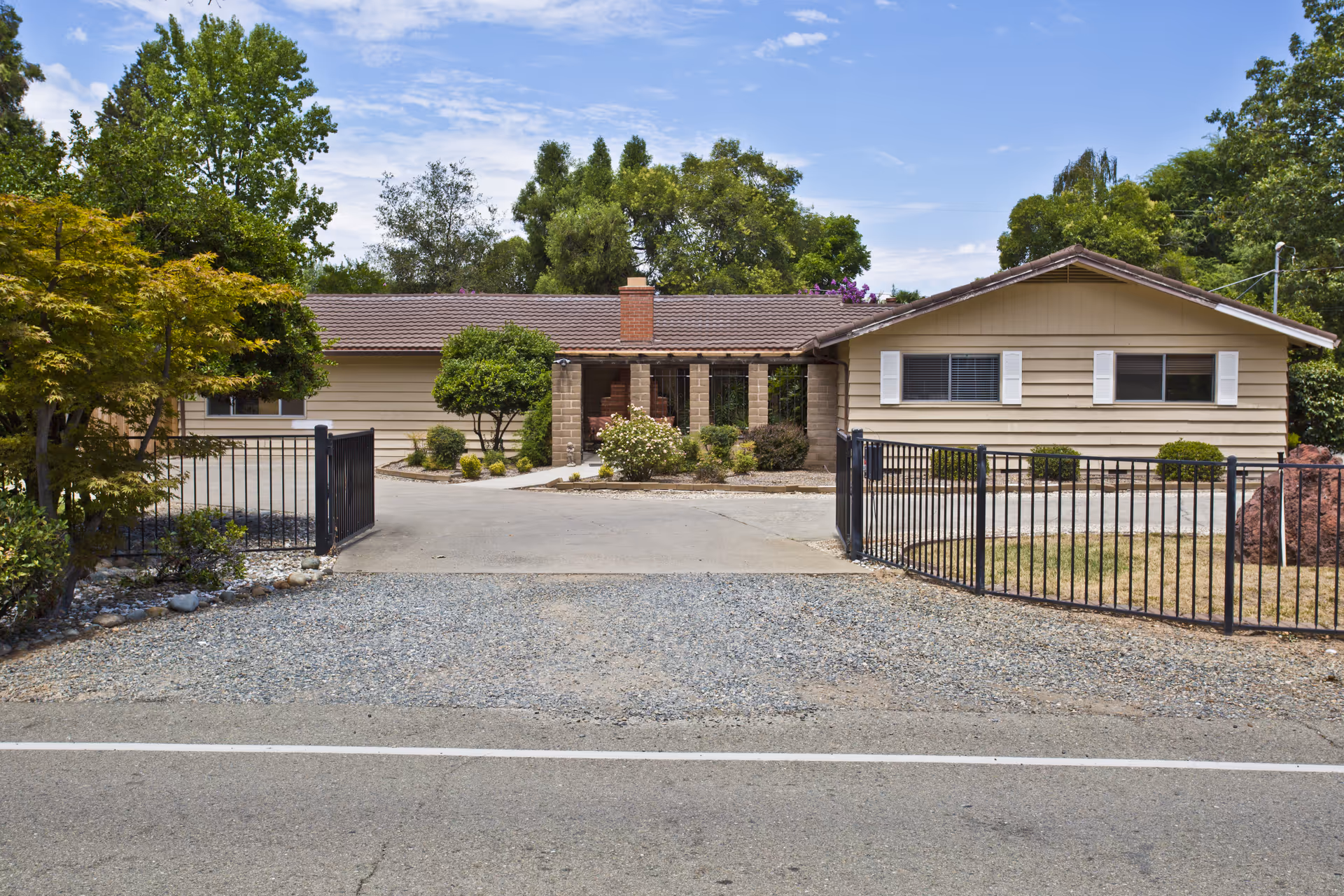 Front exterior view of a single-story residential building with beige siding, a brown tiled roof, and a central covered porch with brick pillars. The property is surrounded by trees and shrubs, with a black metal gate at the driveway entrance.