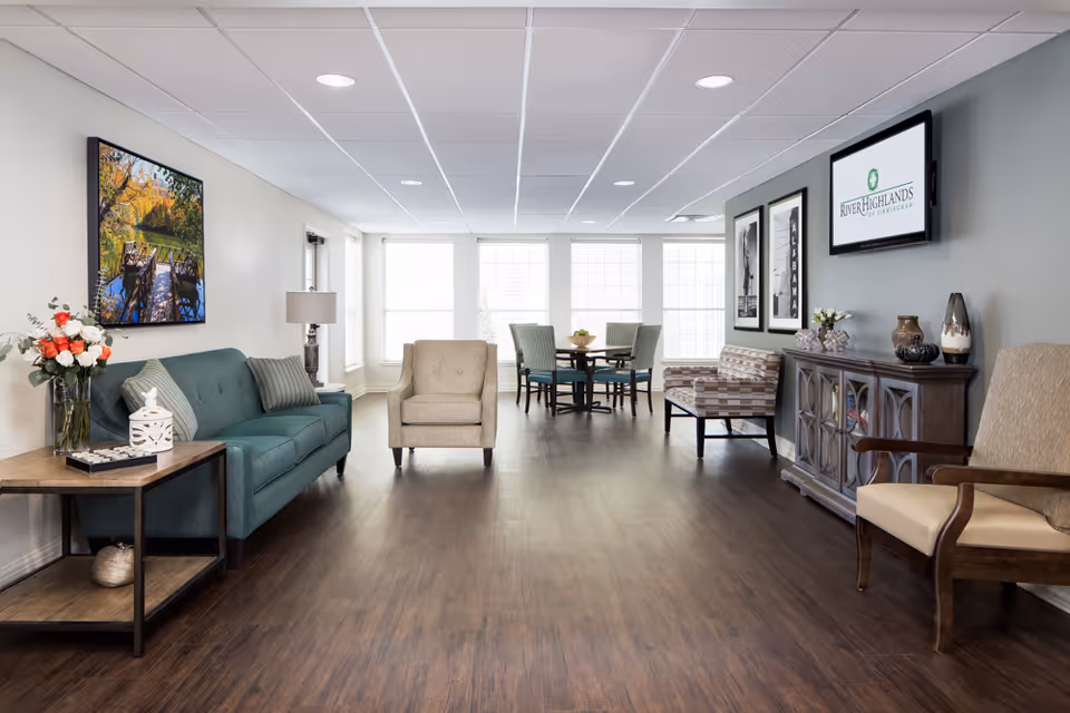 A bright and spacious living room area in a senior living facility featuring a teal sofa with pillows, a beige armchair, a wooden side table with a vase of flowers and decorative items, a patterned bench, and a wooden cabinet with vases and decor. A flat-screen TV mounted on the wall displays the River Highlands of Birmingham logo. Large windows allow natural light to fill the room, and a small dining table with four chairs is visible in the background.