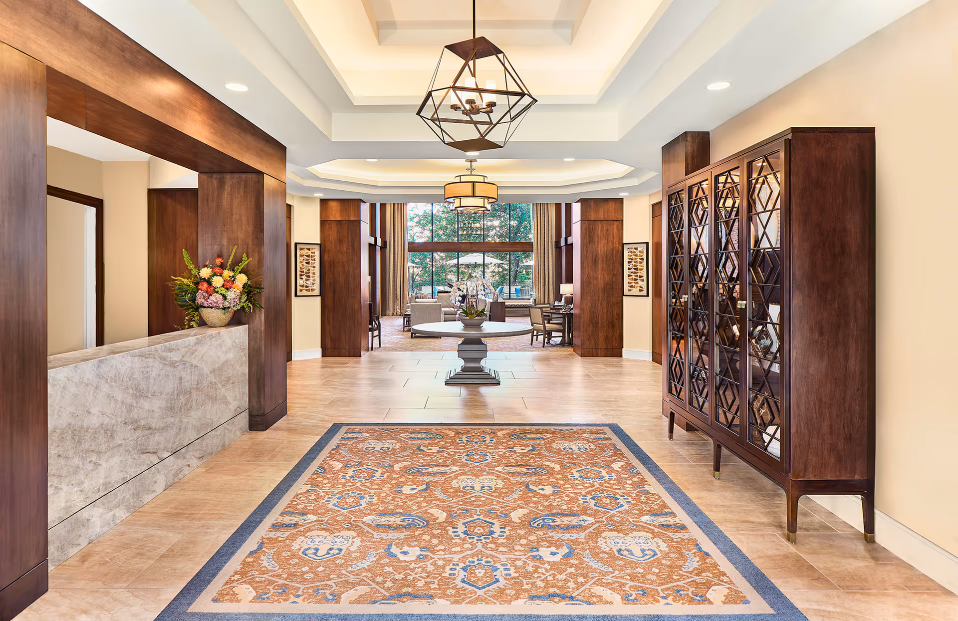 Spacious and elegant interior lobby area with a large patterned rug on the floor, a round table with a flower arrangement in the center, wooden pillars and cabinetry, modern pendant lighting, and large windows showing an outdoor seating area with greenery.