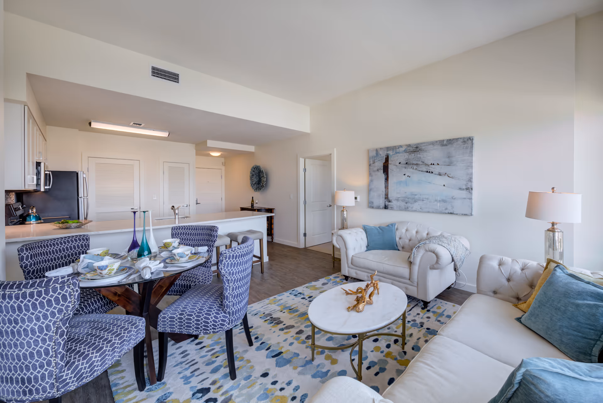 A bright and modern living and dining area in a senior living facility. The space features a glass dining table with four patterned blue chairs, a kitchen with white cabinets and stainless steel appliances, and a cozy living room with two white tufted sofas, blue and green cushions, a round marble coffee table, and a colorful patterned rug. A large abstract painting hangs on the wall, and two table lamps provide additional lighting.