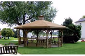 A wooden gazebo with a shingled roof situated on a grassy lawn surrounded by trees and shrubs. There are benches inside and outside the gazebo, and a building is partially visible in the background.