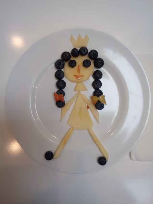 A white plate with a creative food art figure made from fruit pieces. The figure resembles a person with a crown, made from slices of apple and blueberries for hair and eyes, with small pieces of other fruit for details like a nose, mouth, and bows on the hair.