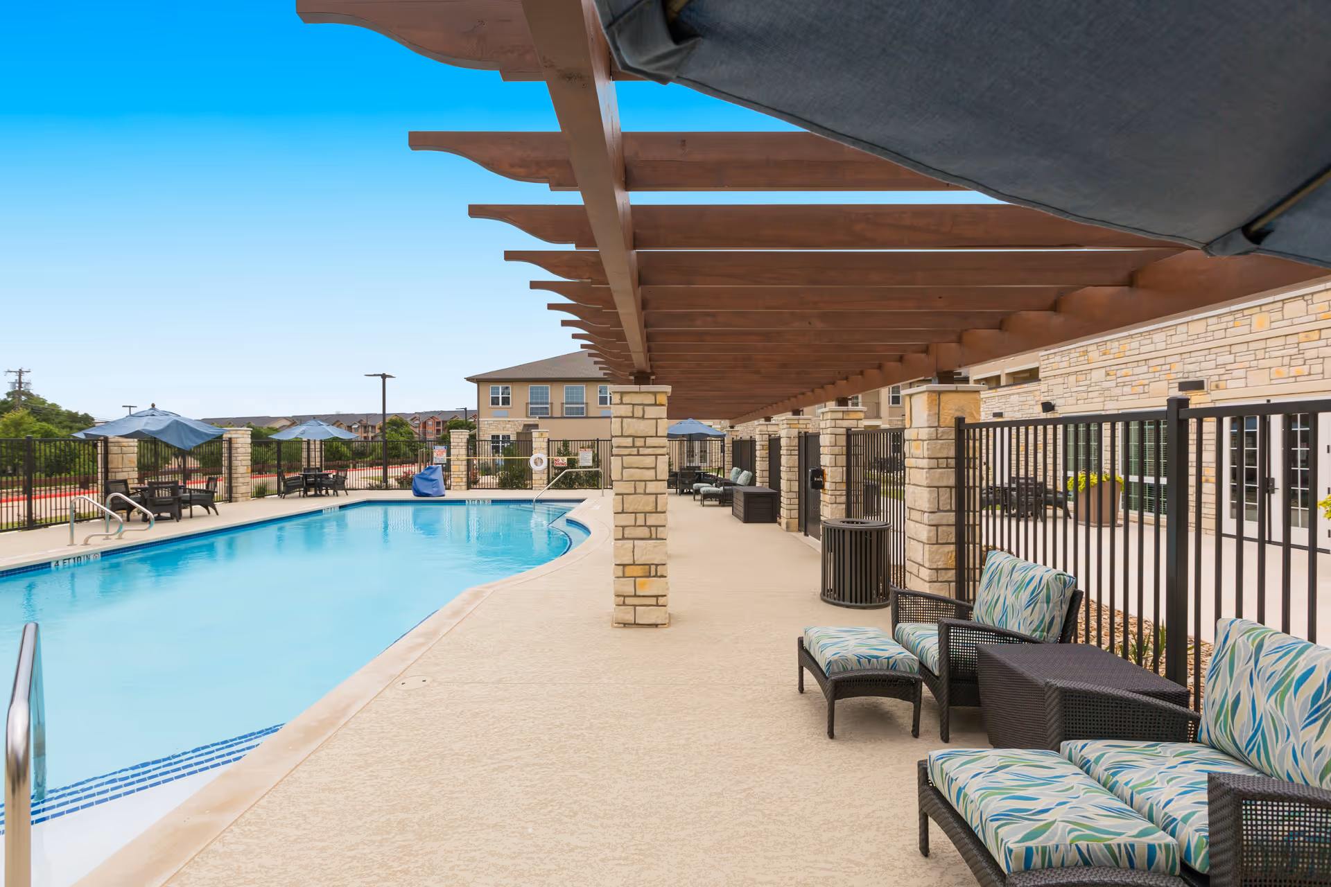 Outdoor swimming pool area with lounge chairs and umbrellas. There is a wooden pergola providing shade over a seating area with cushioned chairs and a small table. The pool is surrounded by a fence, and a building is visible in the background under a clear blue sky.