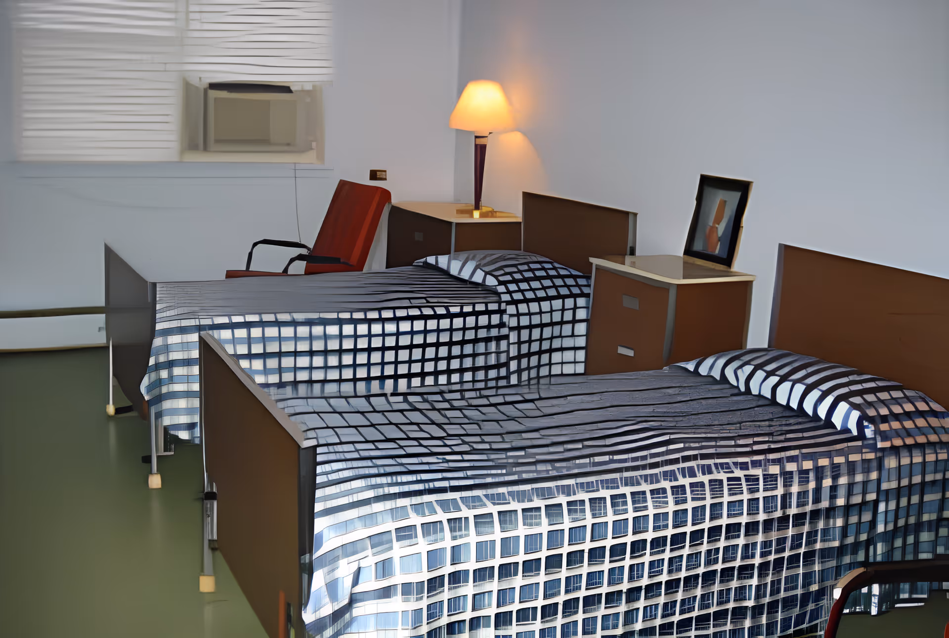 A simple bedroom with two single beds covered in blue and white checkered bedspreads. Between the beds is a small nightstand with a framed picture on it. A lamp on a corner table provides warm light. There is a red chair near a window with blinds and an air conditioning unit.