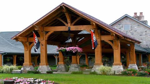 Entrance of The Inn at CoalRidge featuring a large wooden covered porch with stone pillars, hanging flower baskets, and flags. The building is made of stone with a shingled roof and surrounded by green grass and flower beds.