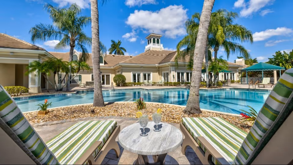 Outdoor pool area at Aston Gardens At Parkland Commons with two striped lounge chairs and a small round table holding two glasses of water with lemon slices. Palm trees and a beige building with a cupola are visible in the background under a blue sky with some clouds.