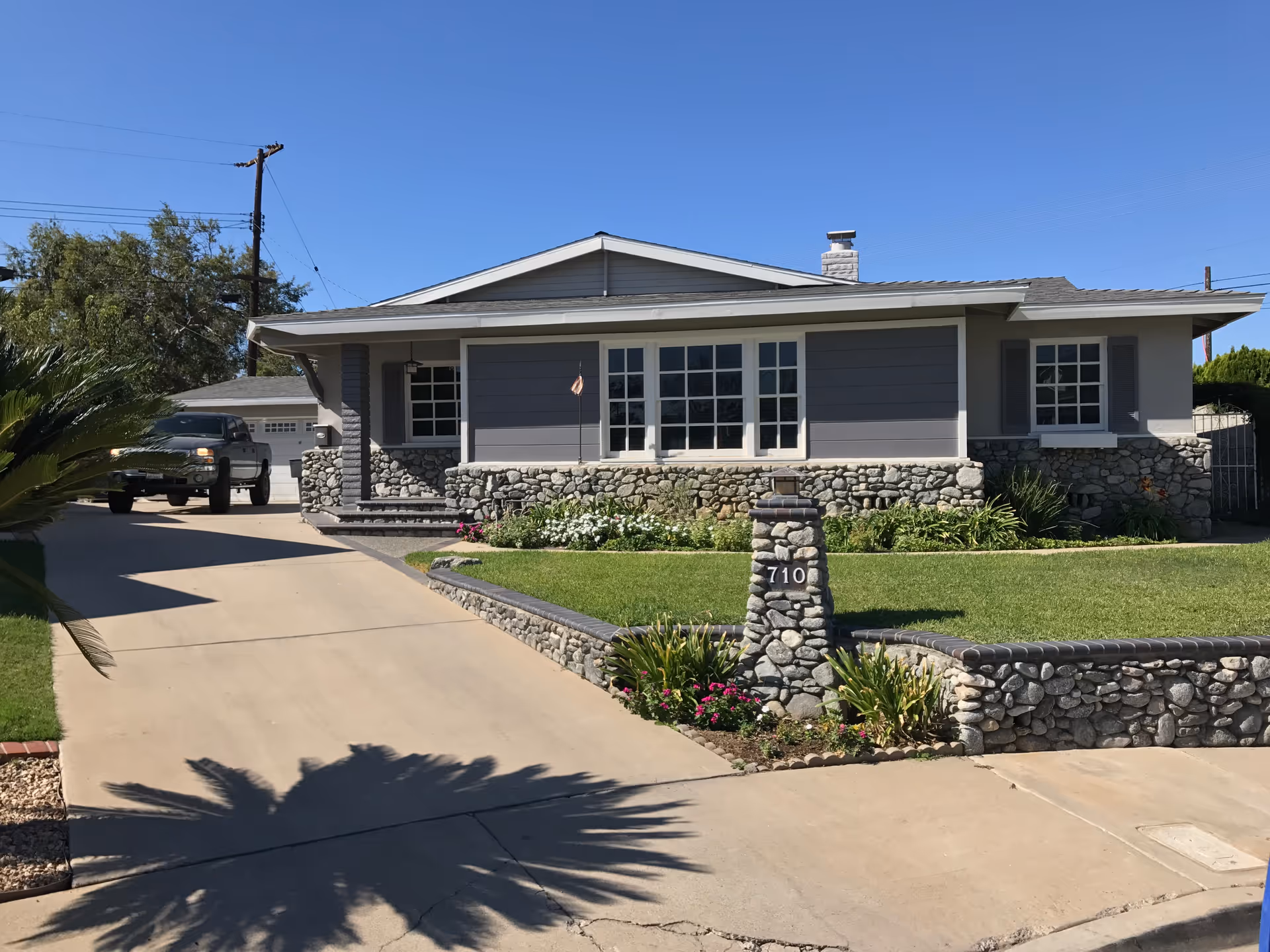 Single-story house with stone and gray siding exterior, a well-maintained front lawn, a stone mailbox with the number 710, and a driveway with a parked pickup truck under a clear blue sky.