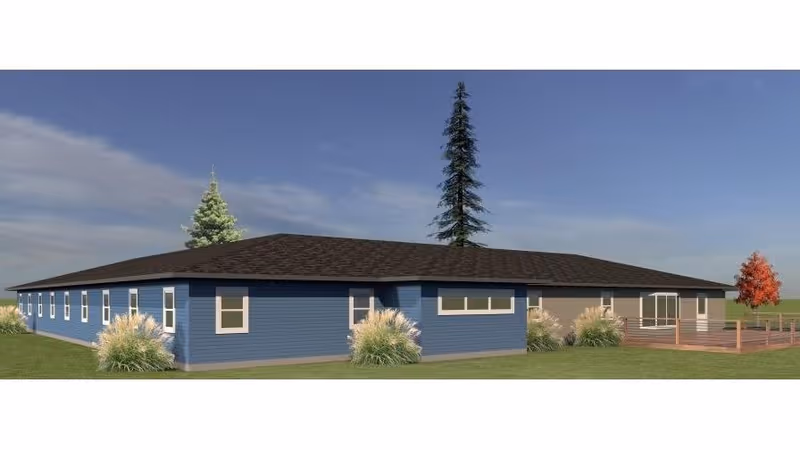 Exterior view of a single-story building with blue and beige siding, multiple windows, a dark shingled roof, surrounded by grass and a few trees under a partly cloudy sky.