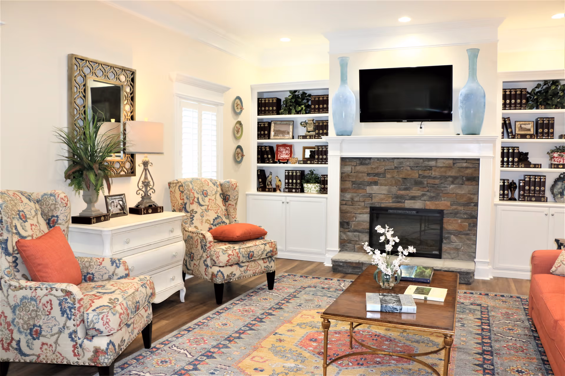 A cozy living room with two patterned armchairs with red cushions, a white side table with a lamp and plant, a wooden coffee table with books and a flower vase, a stone fireplace with a mounted flat-screen TV above it, and built-in white shelves filled with books and decorative items.