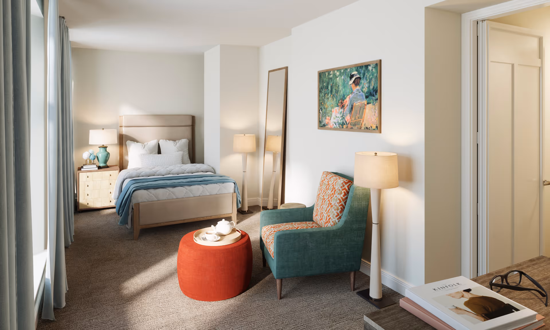 A bright and cozy bedroom with a beige upholstered bed, white and blue bedding, a wooden nightstand with a green lamp, a tall mirror leaning against the wall, a green armchair with an orange patterned cushion, a red round ottoman with a tea set on a tray, two floor lamps, and a framed painting of a woman sitting in a garden on the wall.
