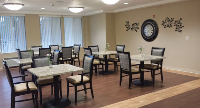 A dining room with several square tables, each surrounded by four chairs with cushioned seats and backs. Each table has a small vase with a flower. The room has wood flooring with a lighter wood border, beige walls, and large windows with vertical blinds. A large round clock and decorative wall art are mounted on one wall.