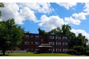 Exterior view of a three-story brick building with multiple windows, surrounded by trees and green grass under a partly cloudy blue sky.