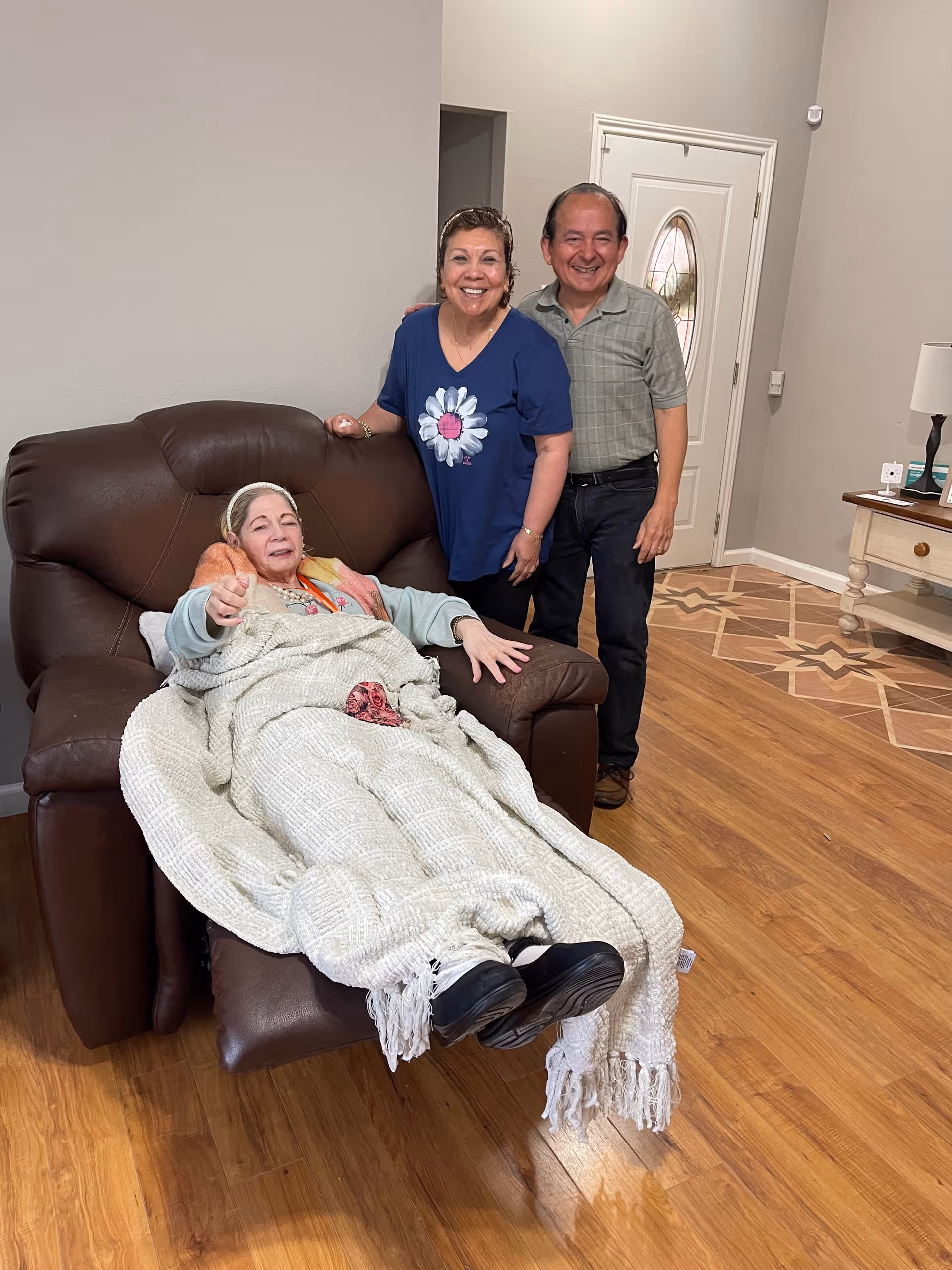 An elderly woman is reclining on a brown leather recliner chair covered with a beige blanket. Behind her, a smiling middle-aged woman in a blue shirt with a flower design and a middle-aged man in a green plaid shirt stand close together in a warmly lit room with wooden flooring and a patterned tile area near the door.
