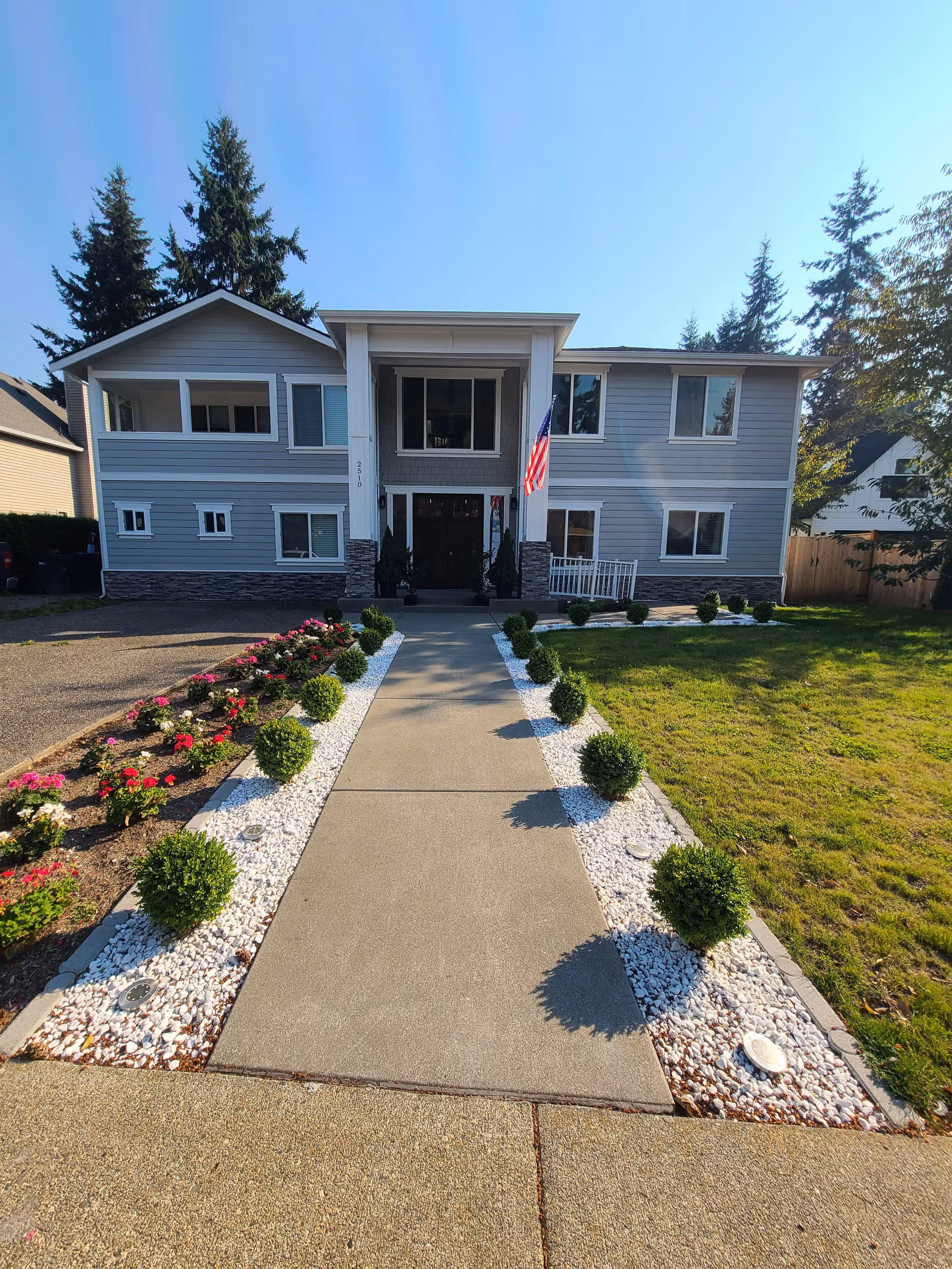 Front exterior view of a two-story senior care facility building with gray siding and stone accents. A concrete walkway lined with small round bushes and white decorative stones leads to the main entrance. There are flower beds with red and white flowers on the left side and a green lawn on the right. An American flag is displayed near the entrance under a covered porch. Tall evergreen trees are visible in the background under a clear blue sky.