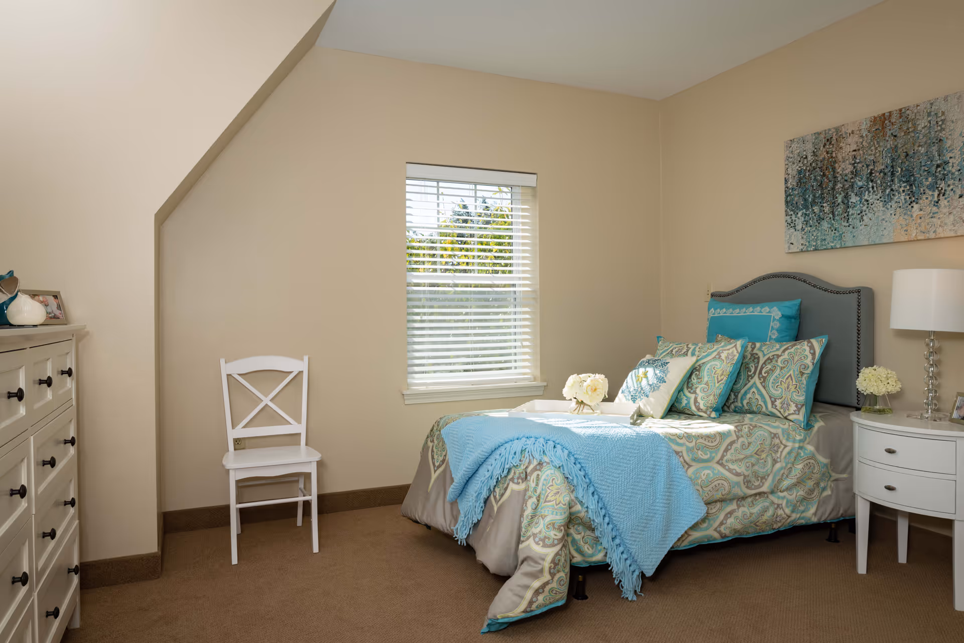 A cozy bedroom with a single bed adorned with patterned bedding and multiple pillows in shades of blue and beige. A light blue throw blanket is draped over the bed. Next to the bed is a white nightstand with a lamp and a small flower arrangement. A white chair is placed against the beige wall near a window with white blinds, allowing natural light to brighten the room. A dresser with black knobs and decorative items is visible on the left side of the room. A large abstract painting in blue tones hangs above the bed.