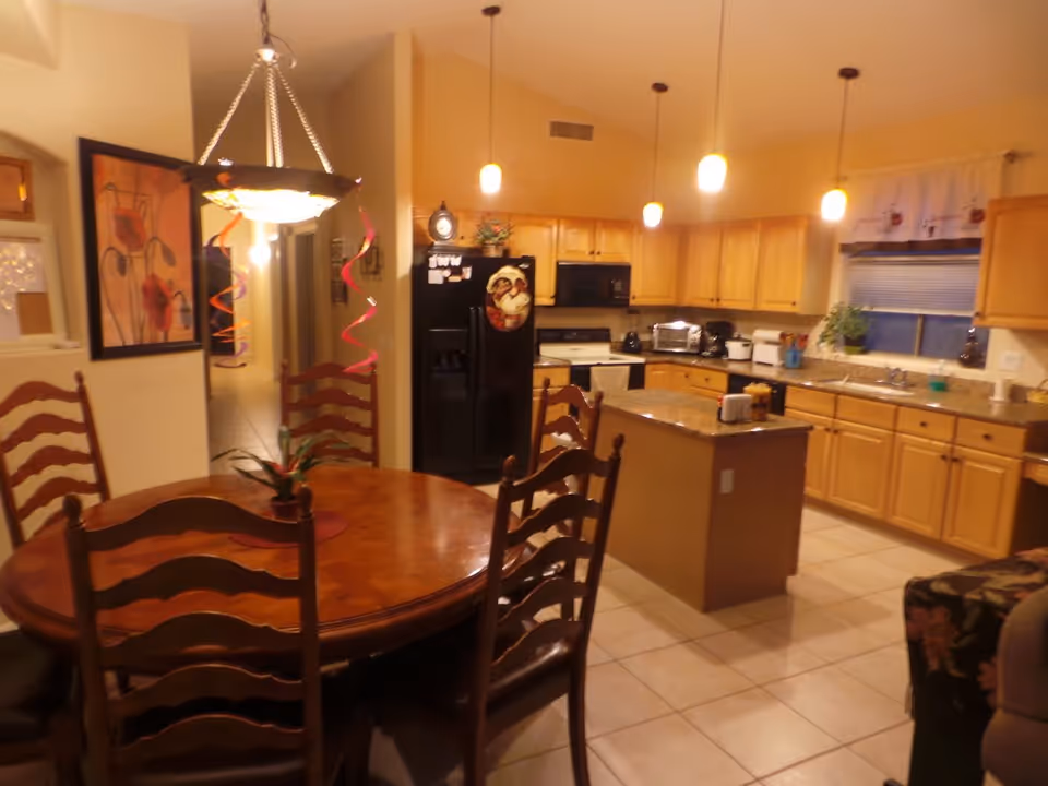 Interior view of a kitchen and dining area in a senior care home. The dining area features a round wooden table with six matching chairs. Above the table hangs a decorative light fixture with red spiral ribbons. The kitchen has light wood cabinets, a black refrigerator, a stove, a microwave, and a central island with a granite countertop. The floor is tiled, and there is a window with a white curtain above the sink. Various kitchen appliances and plants are visible on the countertops.