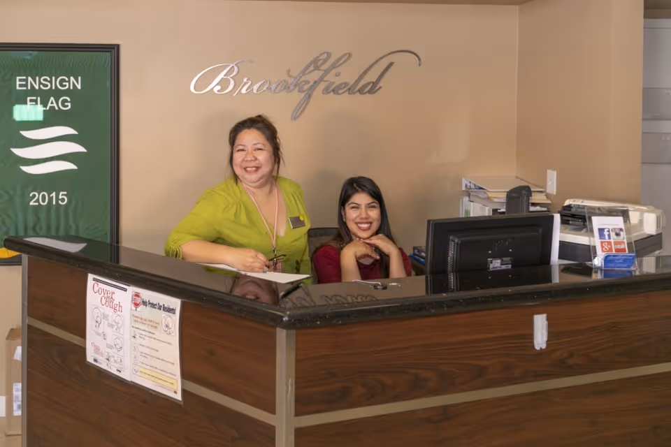 Two smiling women at a reception desk inside Brookfield Healthcare Center. One woman is standing and wearing a green top, while the other is seated and wearing a maroon top. Behind them on the wall is a sign that says 'Brookfield'. There is also a green banner on the left side with the text 'ENSIGN FLAG 2015'.