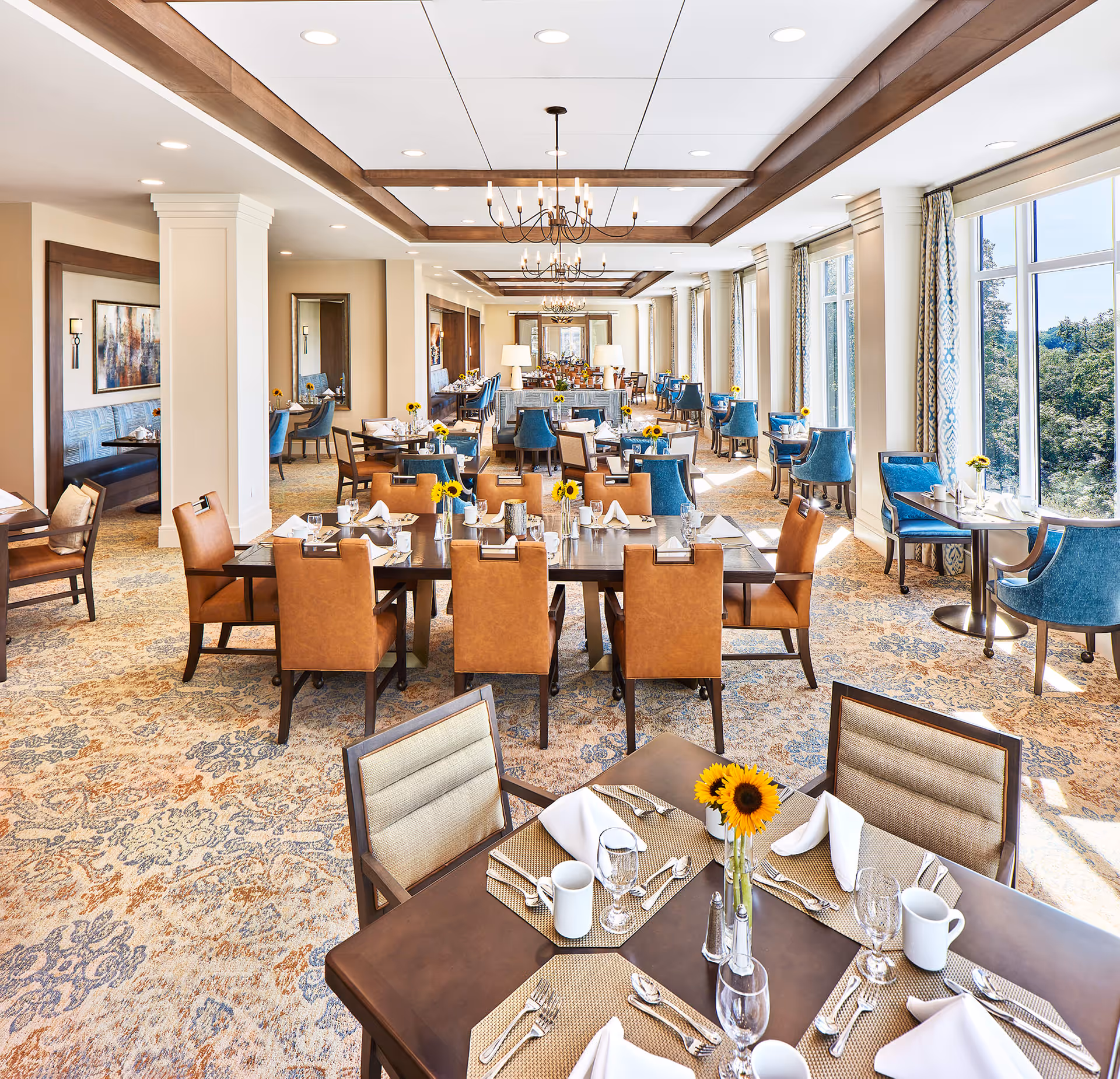 Bright and spacious dining room with multiple tables set for a meal. Tables have brown and blue chairs, white napkins, silverware, glassware, and small vases with sunflowers. Large windows on the right side let in natural light and offer a view of greenery outside. The ceiling features wooden beams and chandeliers, and the floor is covered with a patterned carpet.