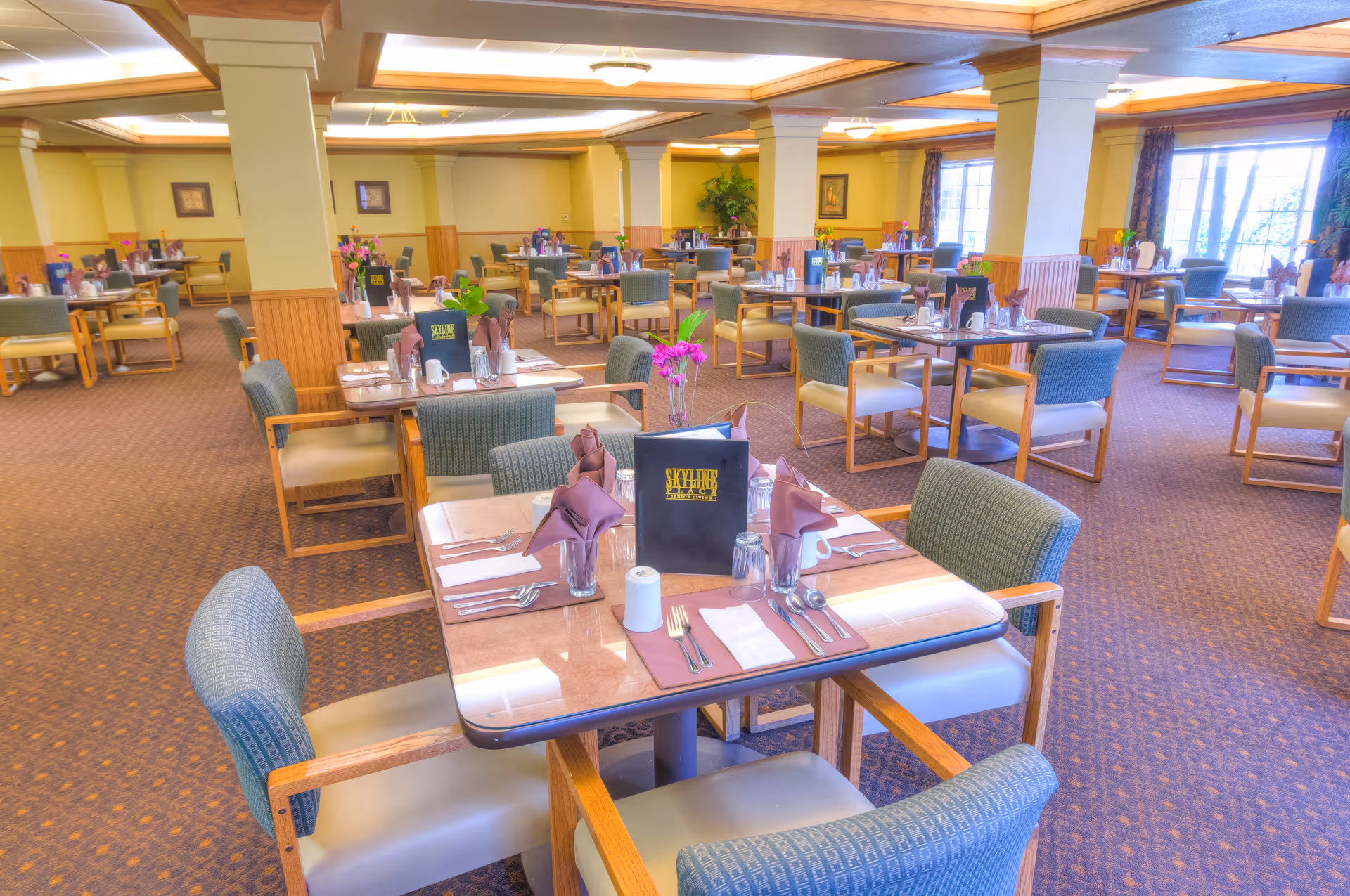 A spacious dining room in a senior living facility with multiple tables set for meals. Each table has chairs with armrests, place settings including utensils, napkins, glasses, and menus labeled Skyline Place Senior Living. The room has carpeted floors, large windows letting in natural light, and decorative plants and flowers on some tables.