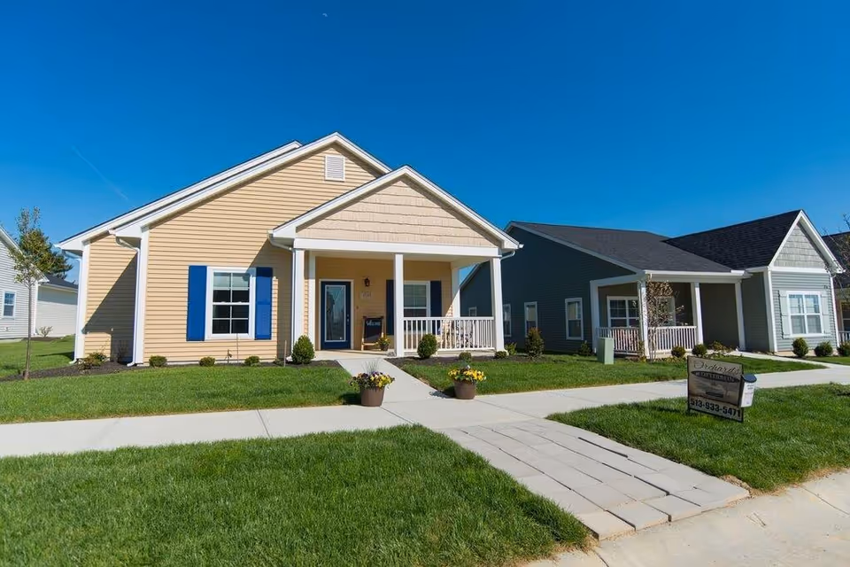 Exterior view of two single-story houses with front porches, well-maintained lawns, and clear blue sky. The house on the left is beige with blue shutters, and the house on the right is gray with white trim. There is a sidewalk and a small paved path leading to the porch of the beige house. A sign with contact information is placed on the lawn near the gray house.