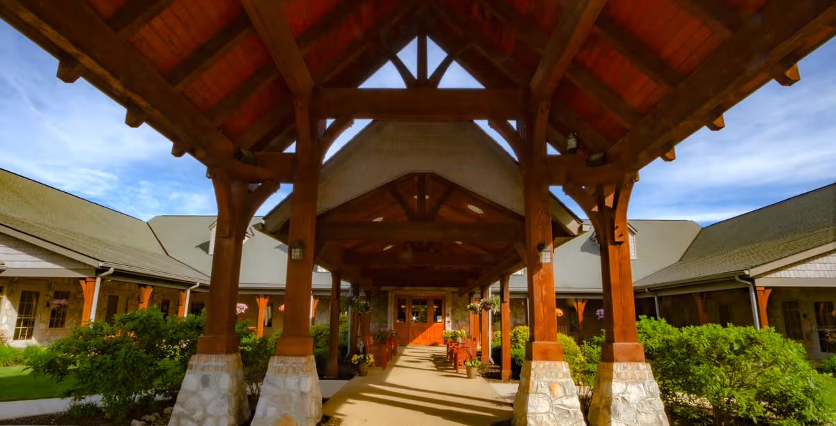 Covered wooden entrance walkway with stone pillars leading to the front doors of a building with a gray roof and beige siding, surrounded by green bushes and hanging flower baskets under a blue sky.