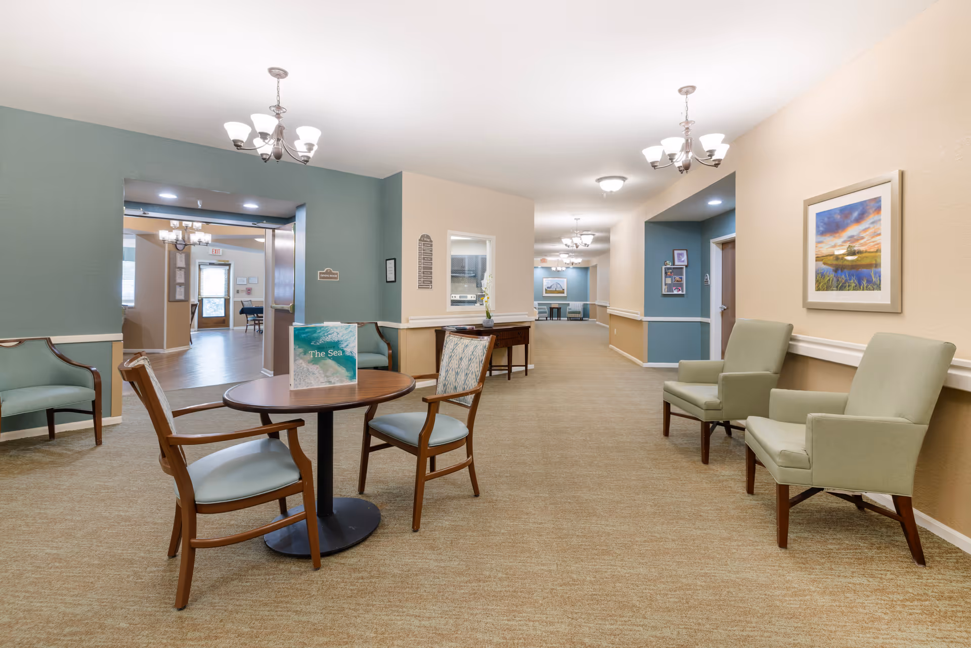 A well-lit hallway in a senior living facility with beige and teal walls, carpeted floor, and several seating areas including wooden chairs with cushions and armchairs. There are chandeliers hanging from the ceiling and framed artwork on the walls. A round table with two chairs is in the foreground, and an open doorway leads to another room in the background.