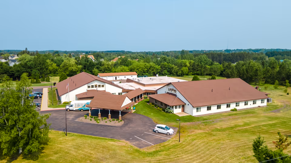Aerial view of a senior living facility named Gardenview surrounded by green lawns and trees. The building has multiple sections with brown roofs and white walls. There are a few cars parked in the parking lot near the entrance.