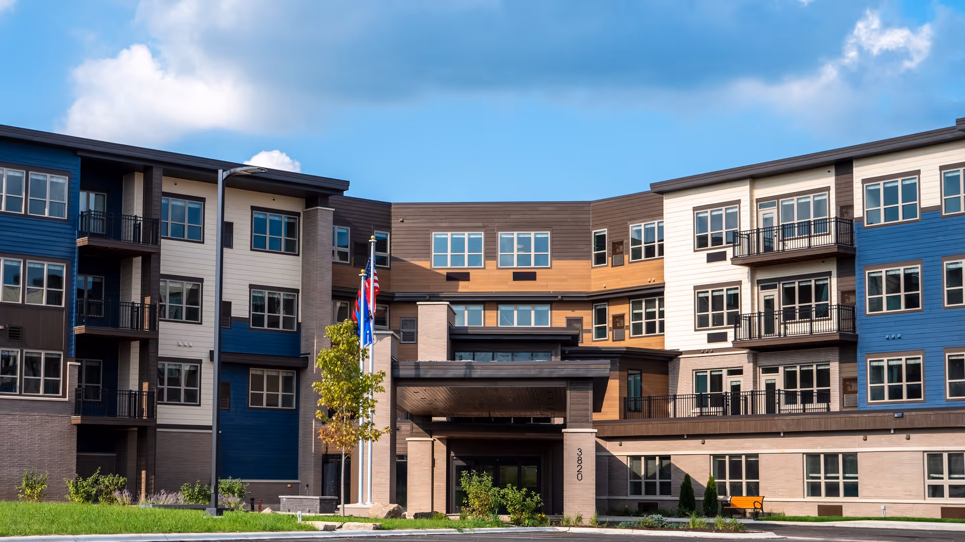 Exterior view of a modern multi-story senior living facility building with a covered entrance, multiple windows, balconies, and three flagpoles with flags in front. The building features a combination of blue, white, and brown siding under a partly cloudy sky.
