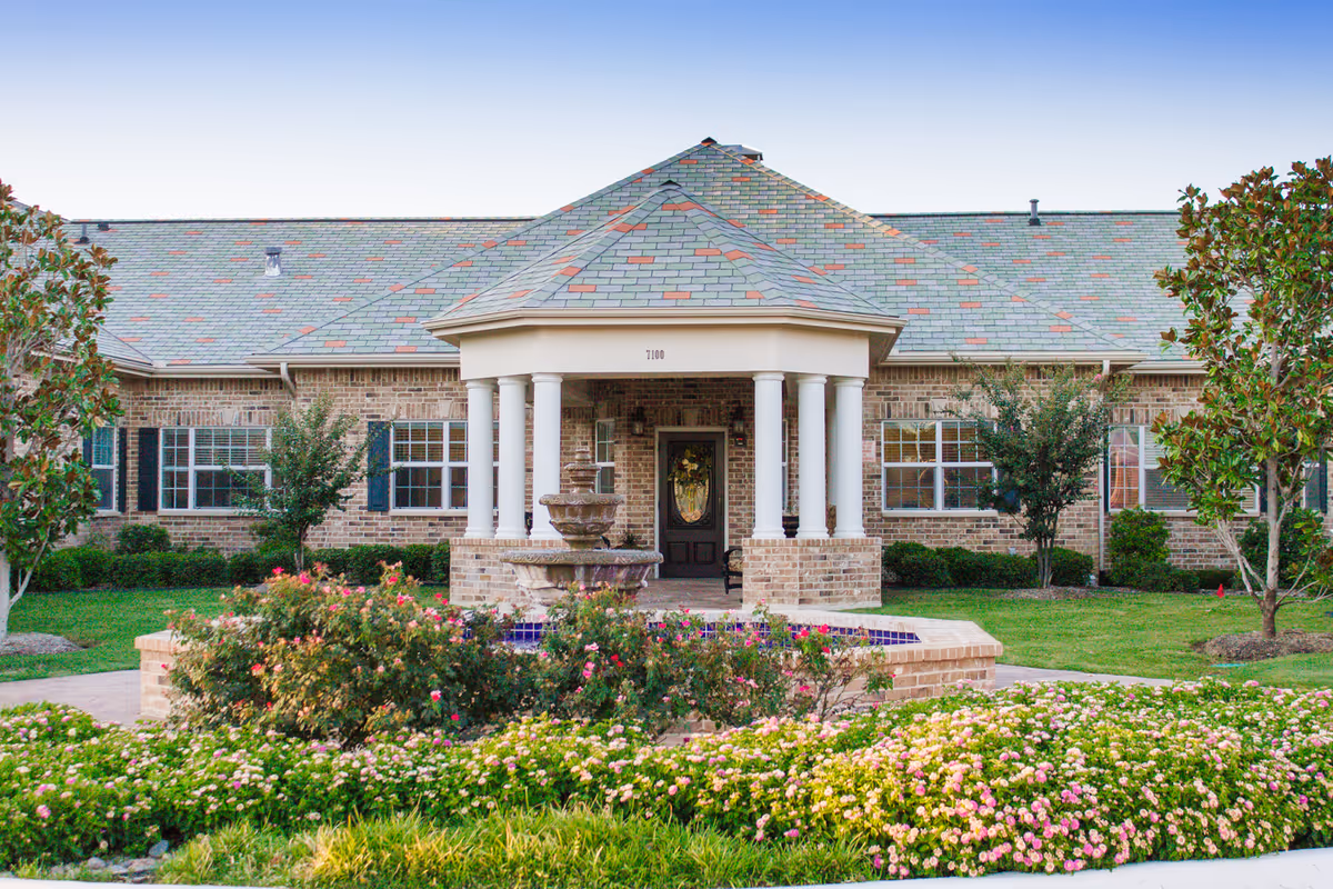 Front exterior view of a single-story brick building with a covered entrance supported by white columns. There is a decorative fountain surrounded by colorful flowers and greenery in the foreground, with trees and shrubs around the building.