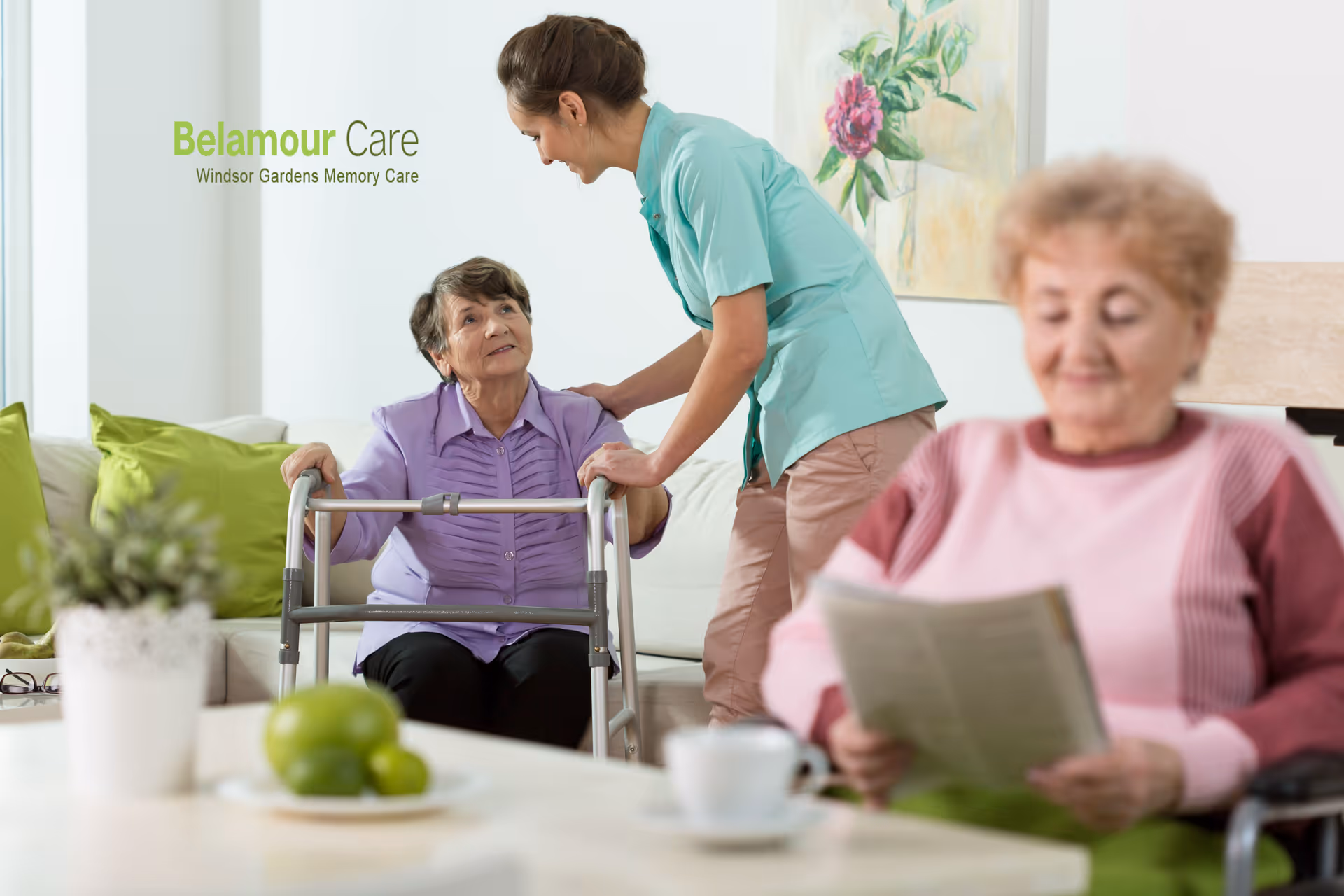 A caregiver in a teal uniform assists an elderly woman using a walker in a bright living room with green pillows and a floral painting on the wall. Another elderly woman in a pink sweater is sitting in the foreground, reading a newspaper.