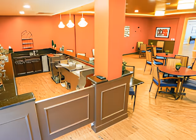 Interior view of a senior living facility dining area with round tables and chairs. The walls are painted orange with brown wainscoting. There is a small kitchen or serving area with countertops and cabinets visible on the left side. Pendant lights hang from the ceiling above the kitchen area.