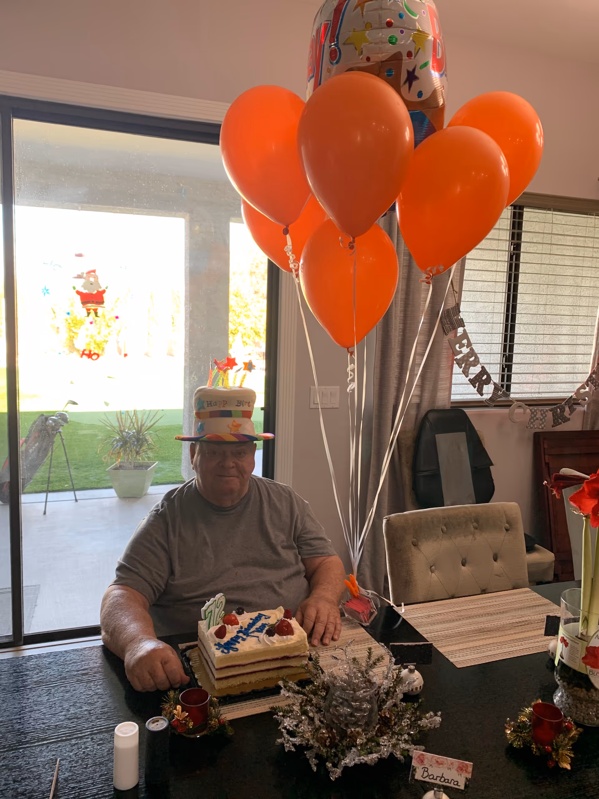 An elderly man sitting at a dining table celebrating his birthday. He is wearing a colorful birthday hat and there are orange balloons tied to the table. A birthday cake with candles and the number 72 is in front of him. The room has a sliding glass door with a view of a green lawn outside and a window with a 'Merry Christmas' banner.