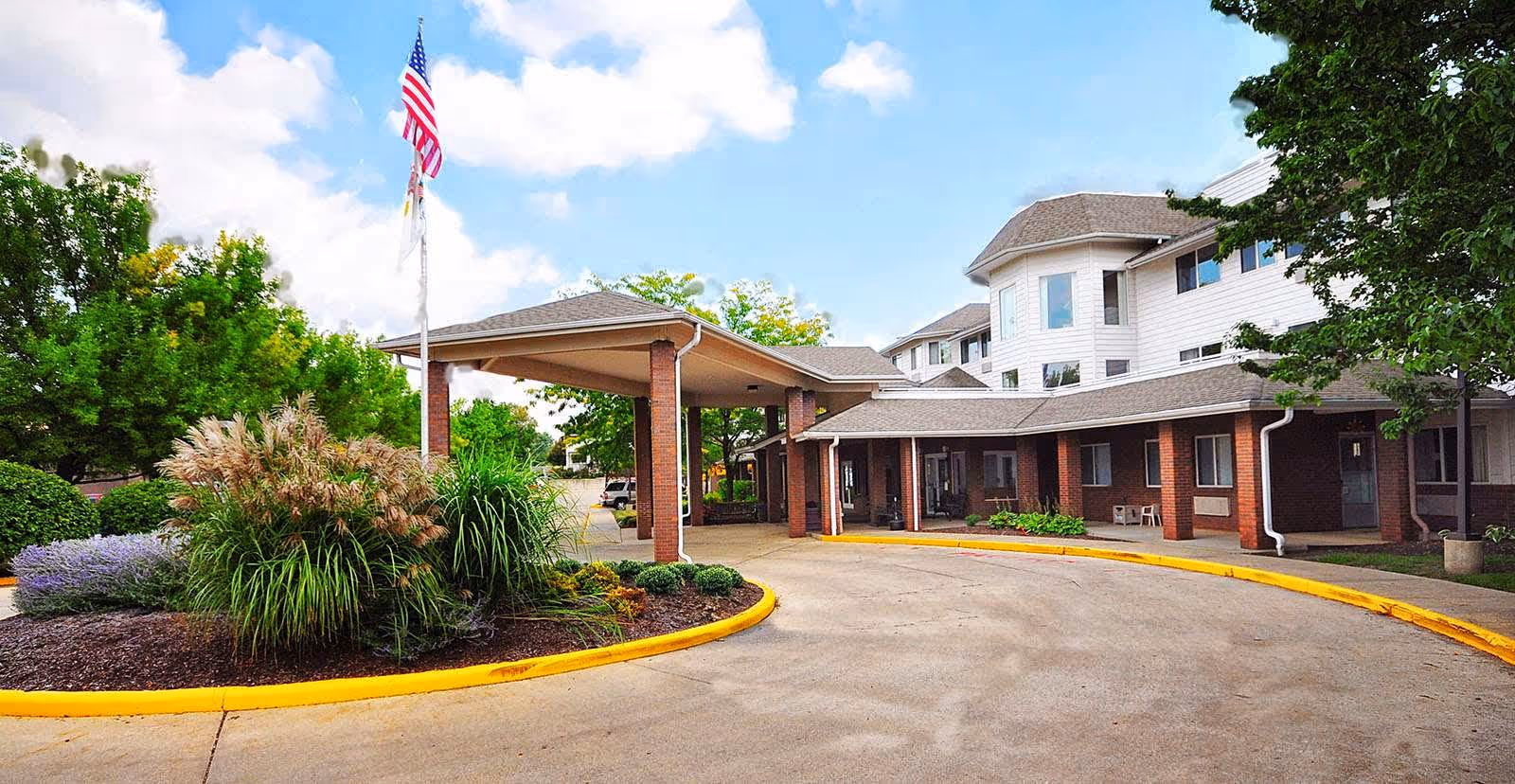 Exterior view of a senior living facility with a covered entrance driveway, landscaped plants, and an American flag on a flagpole. The building has multiple windows and a combination of brick and light-colored siding.