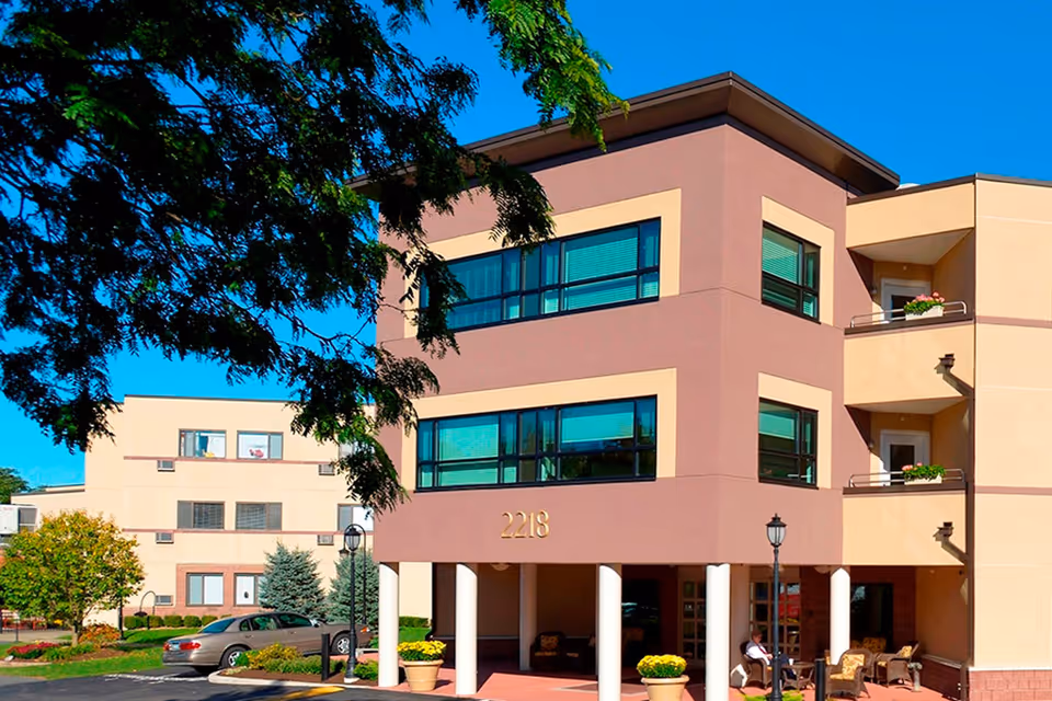 Exterior view of a three-story senior living facility building with beige and brown walls, large windows, balconies with flower pots, and a covered entrance supported by white columns. There are outdoor chairs and tables near the entrance, a tree partially framing the image, and a car parked in the driveway under a clear blue sky.