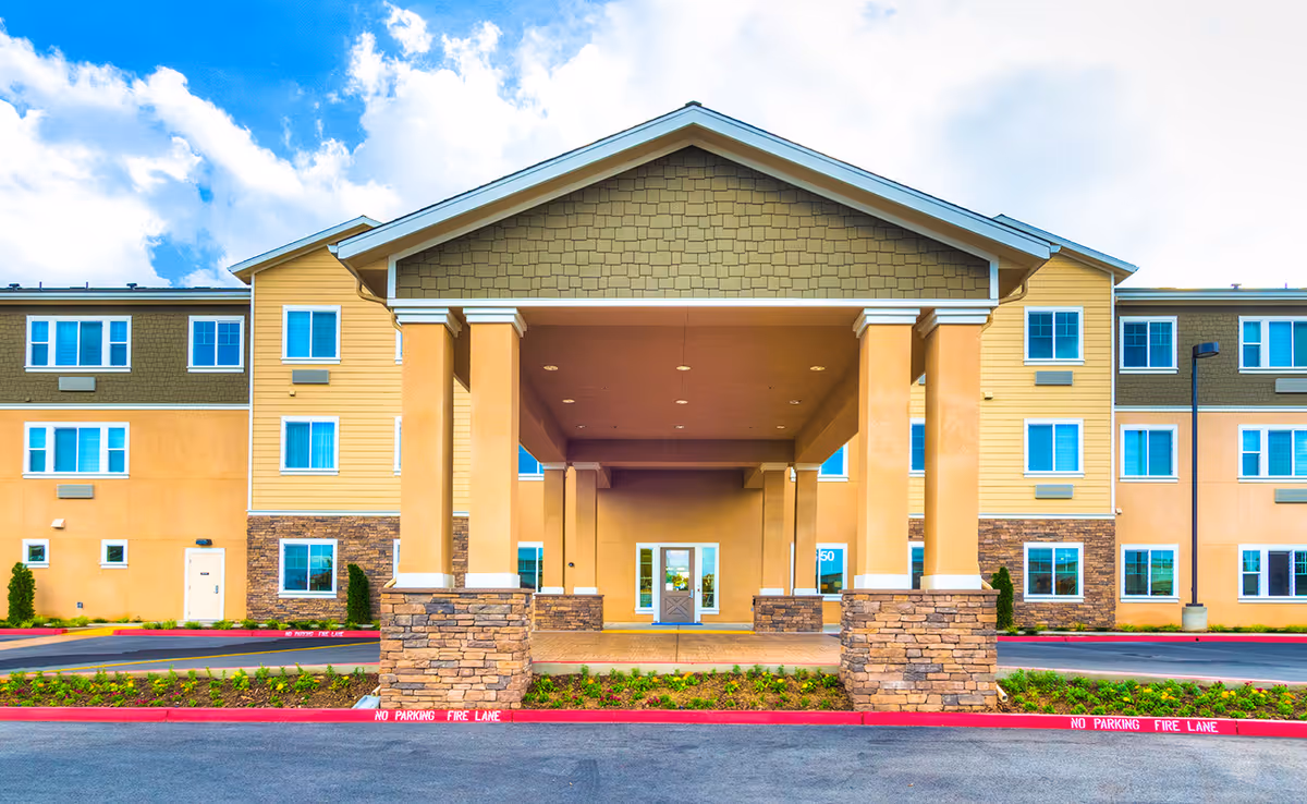 Front exterior view of Carmel Village Memory Care facility showing a large covered entrance with stone pillars, beige and yellow siding, multiple windows, and a clear blue sky with clouds.