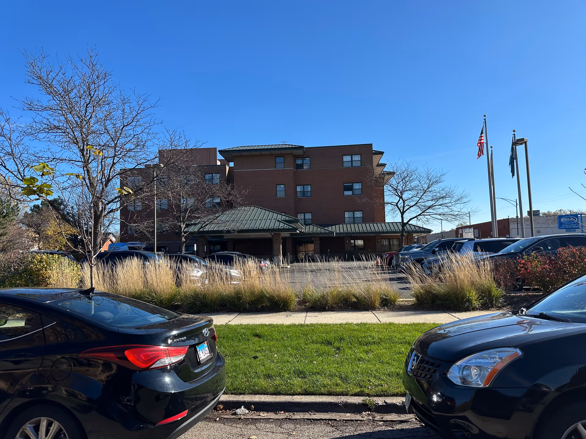 Exterior view of a multi-story brick building with a green roof, surrounded by parked cars and landscaping including grass, bushes, and trees under a clear blue sky. Two flagpoles with flags are visible near the entrance.