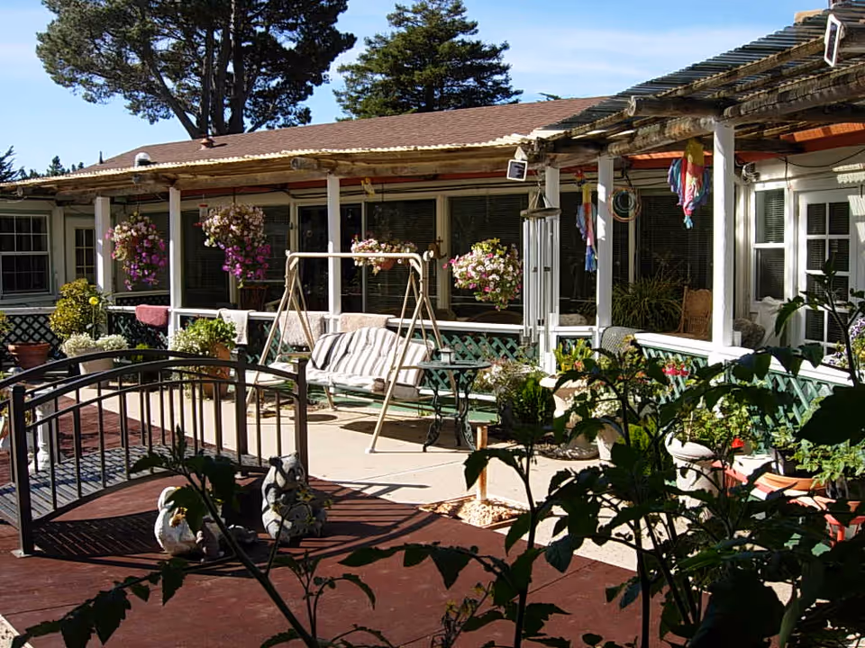 Outdoor patio area at Flanders Court of Carmel featuring a small arched footbridge, garden statues, hanging flower baskets, a cushioned swing bench, potted plants, and a covered porch with seating.