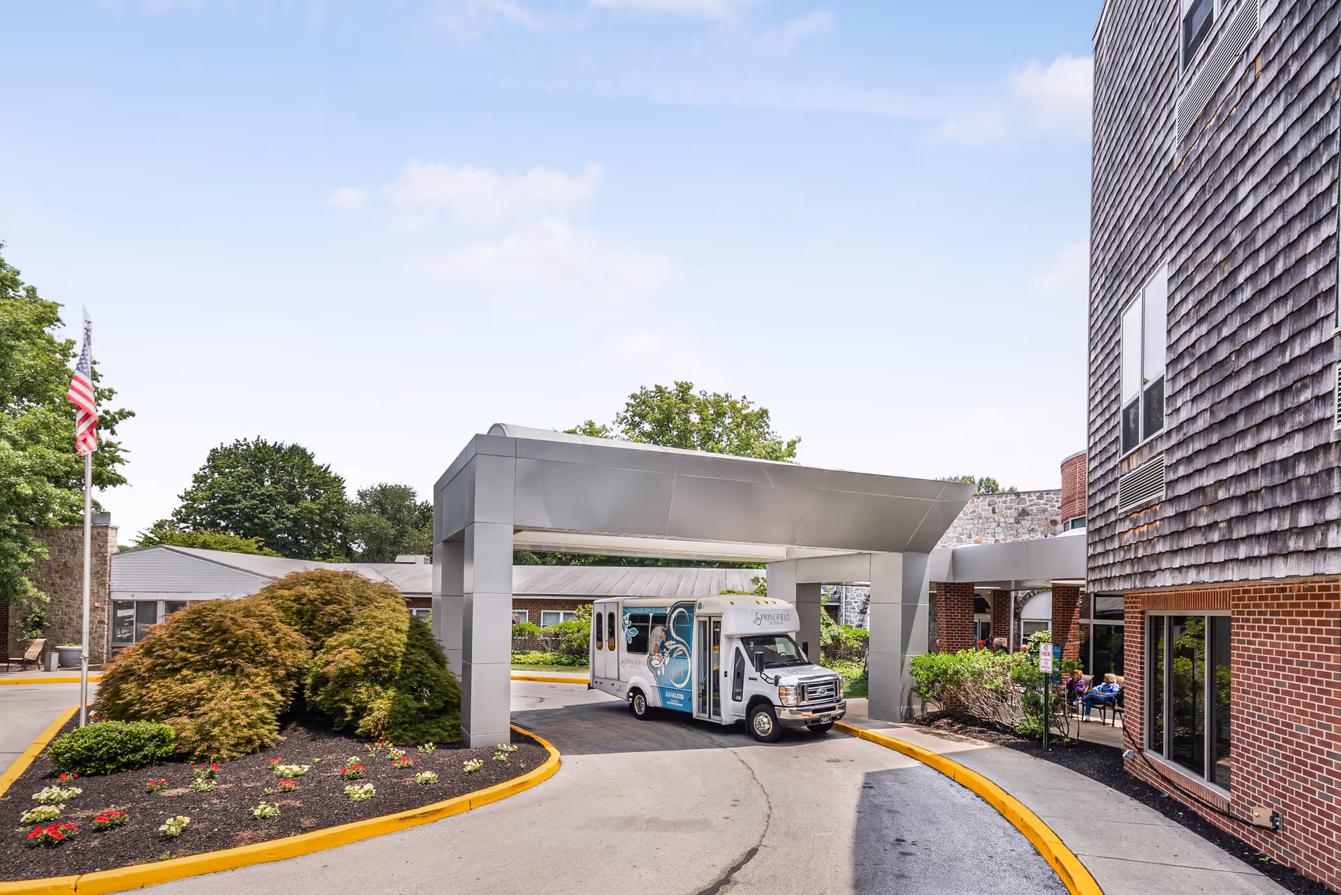 Entrance of Springfield Crossings senior living facility with a covered drop-off area. A small shuttle bus with the Springfield Crossings logo is parked under the canopy. The building exterior features brick and wood siding, with landscaped bushes and flowers near the driveway. An American flag is visible on the left side.
