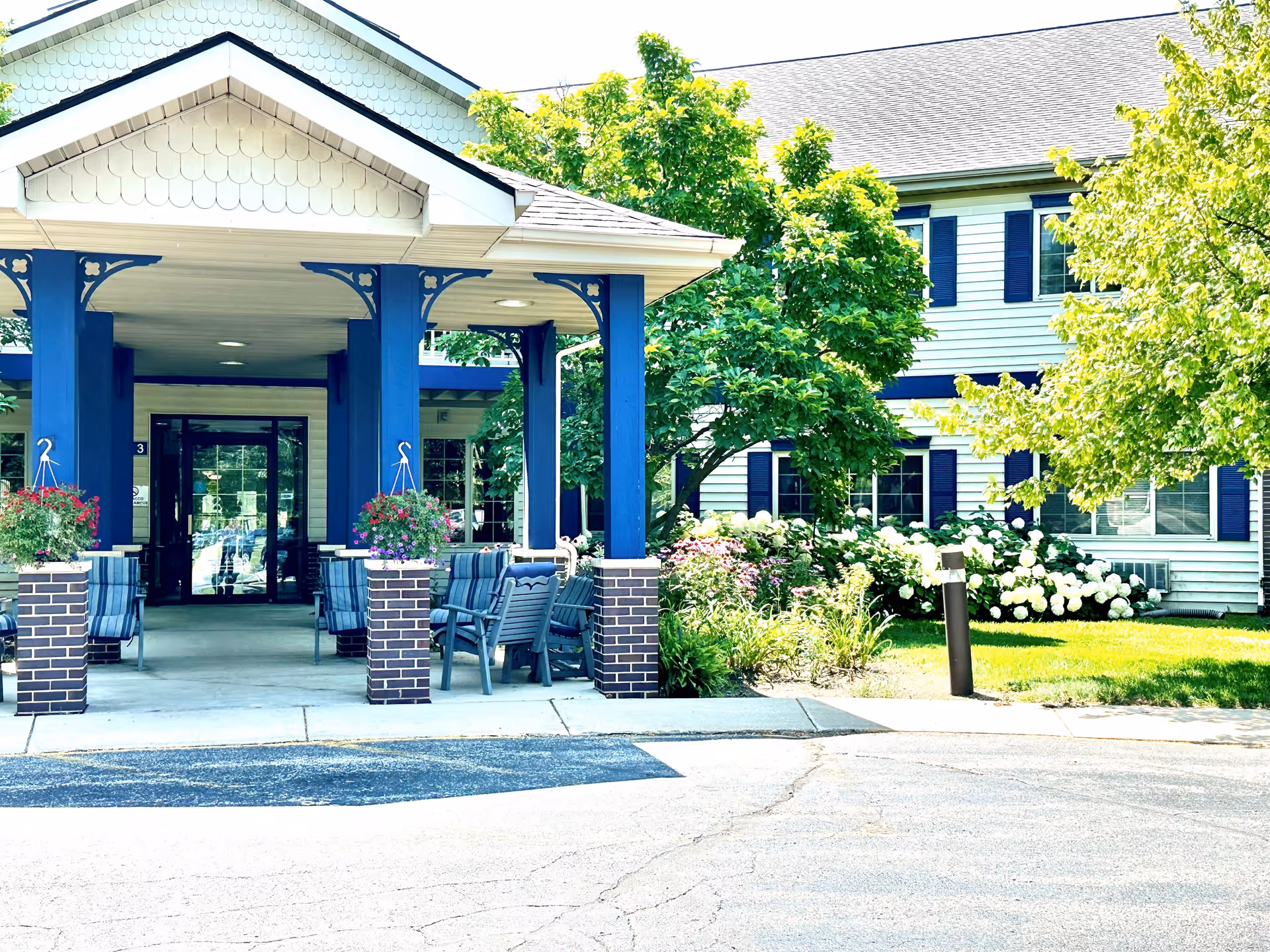 Entrance of a senior living community building with a covered porch supported by blue columns and brick bases. There are several chairs arranged under the porch and hanging flower baskets on the columns. The building has white siding with blue shutters and is surrounded by green trees and flowering bushes.
