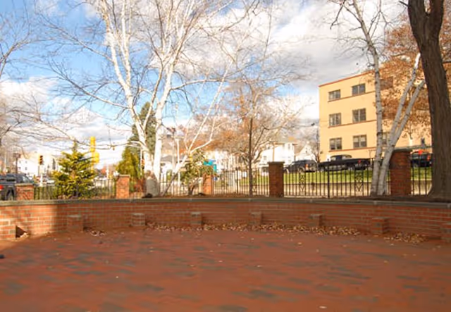 Outdoor brick patio area with a low brick wall and metal railing, surrounded by leafless trees and a multi-story building in the background under a partly cloudy sky.
