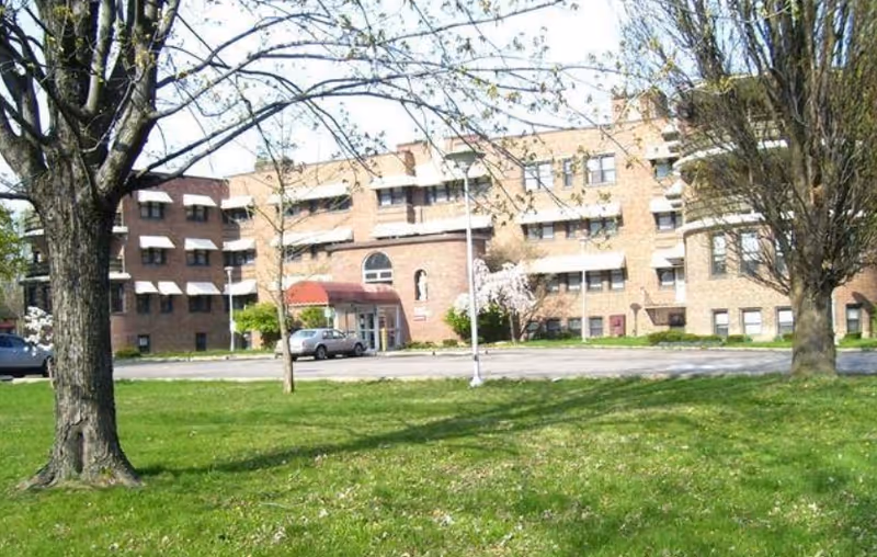 Exterior view of a multi-story brick building with several windows and a red awning over the entrance. The building is surrounded by a grassy lawn with a few trees, some of which have budding leaves. There is a car parked near the entrance and a lamppost on the lawn.