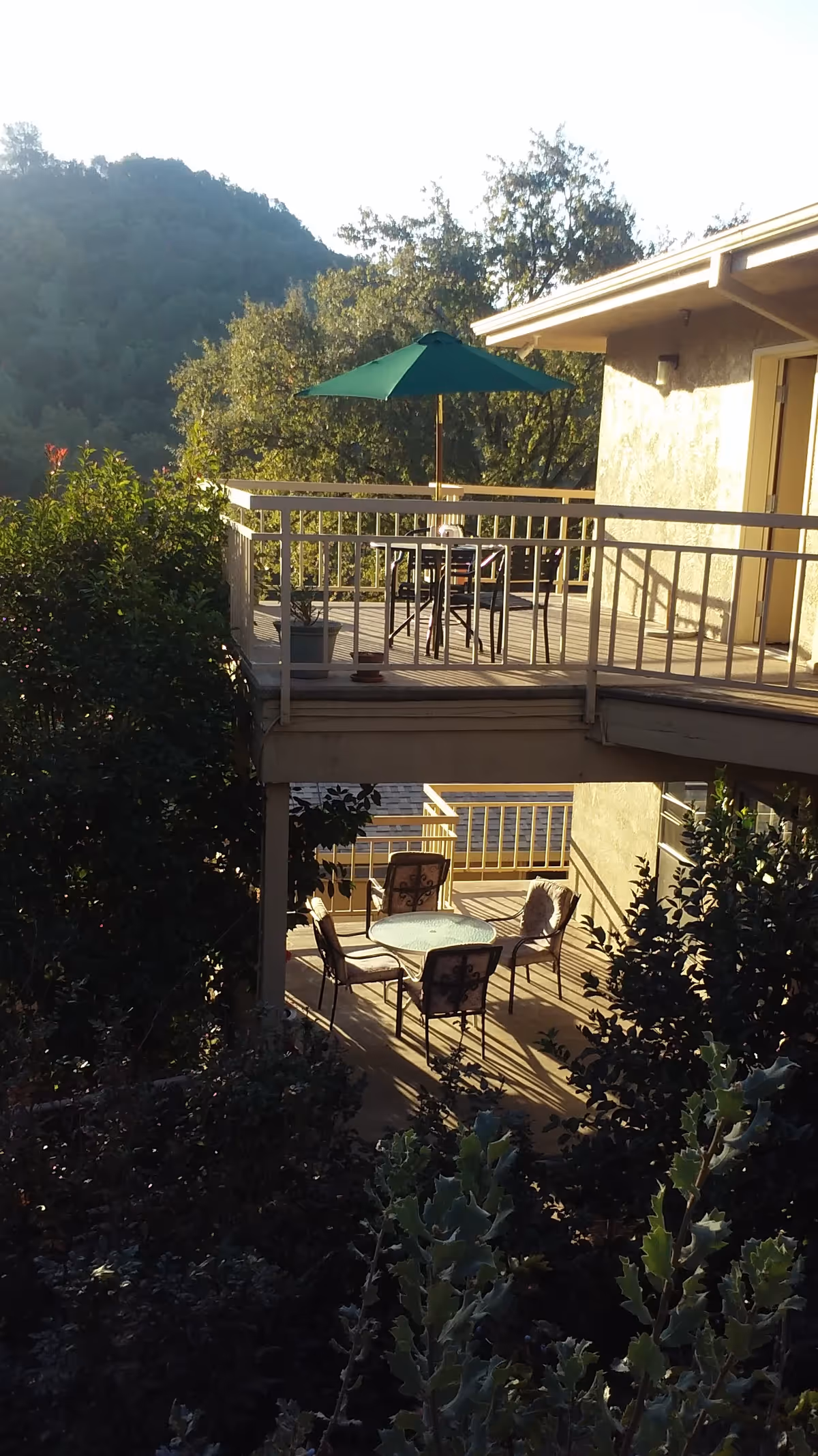 Outdoor patio area of a senior living facility with two levels of wooden decks. The upper deck has a table with chairs and a green umbrella, while the lower deck has a round glass table with four chairs. The area is surrounded by trees and greenery with hills visible in the background.