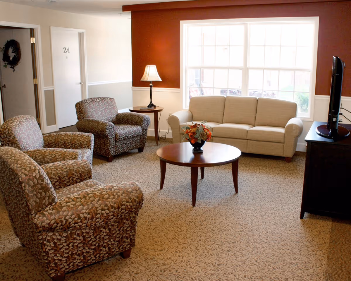 A cozy living room area with a beige sofa, three patterned armchairs, a round wooden coffee table with a floral arrangement, a side table with a lamp, and a flat-screen TV on a dark wooden stand. The room has a large window letting in natural light and walls painted in beige and reddish-brown tones.