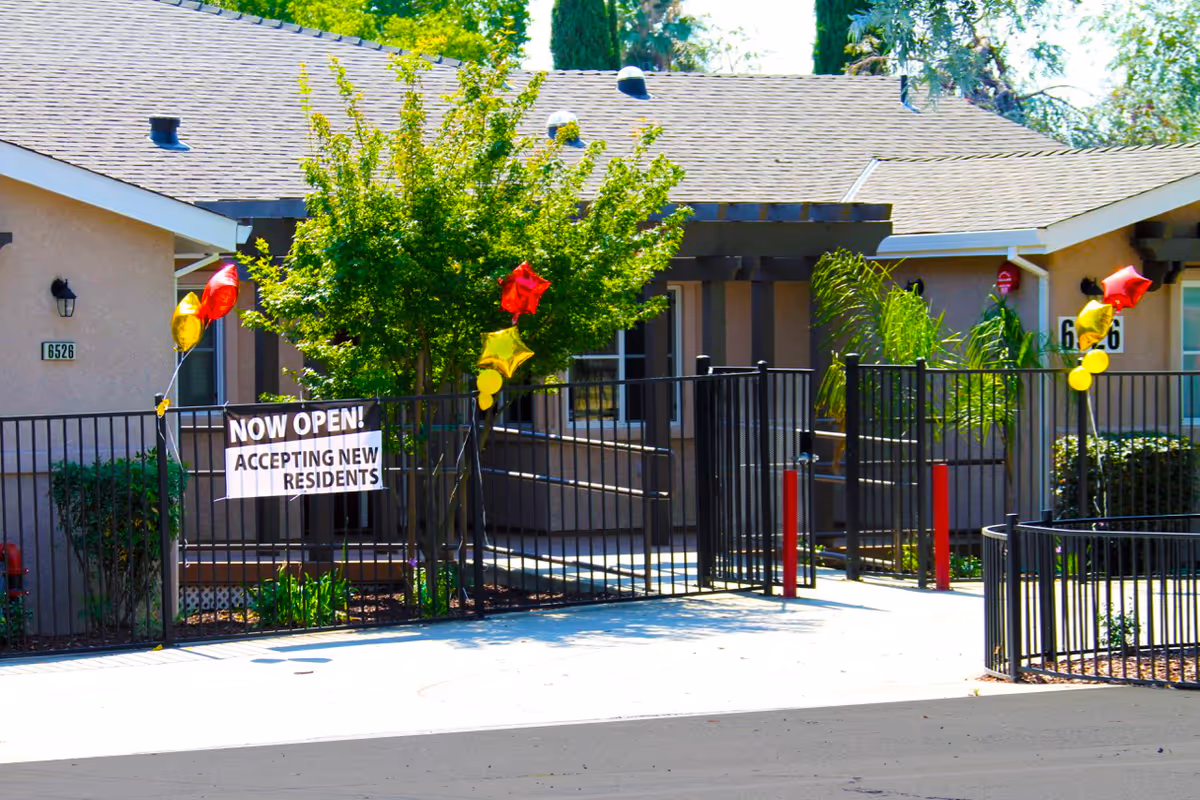 Exterior view of a senior living facility entrance with a black metal fence decorated with red and yellow star-shaped balloons. A sign on the fence reads 'NOW OPEN! ACCEPTING NEW RESIDENTS'. The building has beige walls and a shingled roof, with trees and shrubs around the entrance.