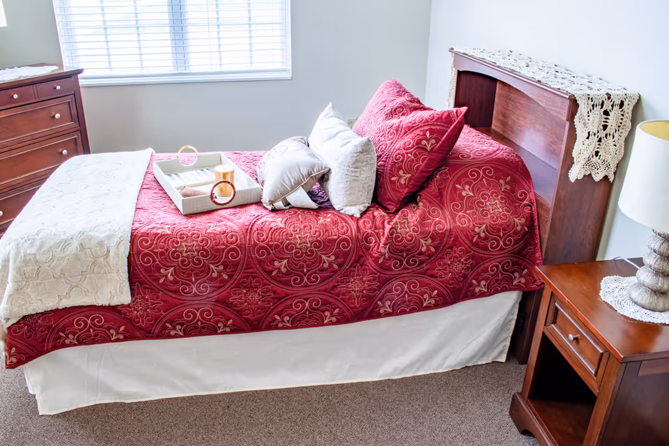 A neatly made single bed with a red patterned bedspread and multiple pillows in a bright room. The bed has a wooden headboard with a white crocheted cloth draped over it. A wooden nightstand with a lamp and a crocheted doily sits beside the bed. A wooden dresser is visible near a window with white blinds.