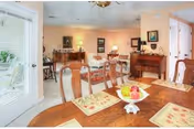 A well-lit dining room with a wooden dining table set with placemats and a fruit bowl centerpiece. The room features multiple wooden chairs, a sideboard with decorative items, framed artwork on the walls, and a glass door leading outside.